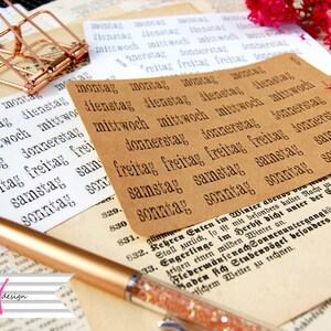 May include: A close-up of a set of brown paper labels with the days of the week written in German: Montag, Dienstag, Mittwoch, Donnerstag, Freitag, Samstag, Sonntag. The labels are arranged in a grid pattern and are surrounded by other papers and a pen.