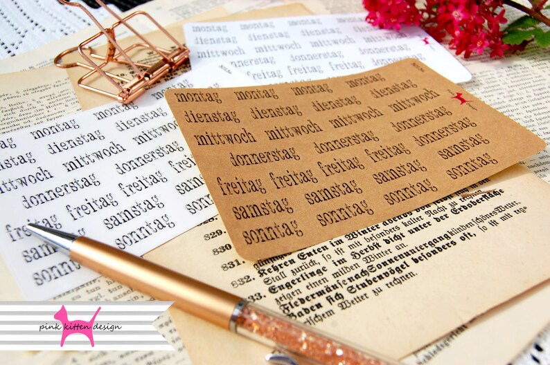 May include: A close-up of a set of brown paper labels with the days of the week written in German: Montag, Dienstag, Mittwoch, Donnerstag, Freitag, Samstag, Sonntag. The labels are arranged in a grid pattern and are surrounded by other papers and a pen.