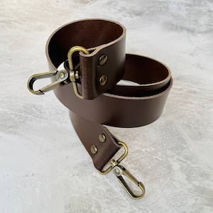 May include: A brown leather strap with two brass clasps. The strap is folded in half and the clasps are visible.