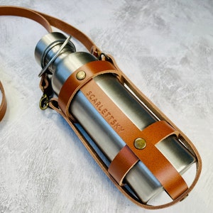 Tan cage style leather water bottle carrier with a crossbody strap and antique brass rivets holding a stainless steel water