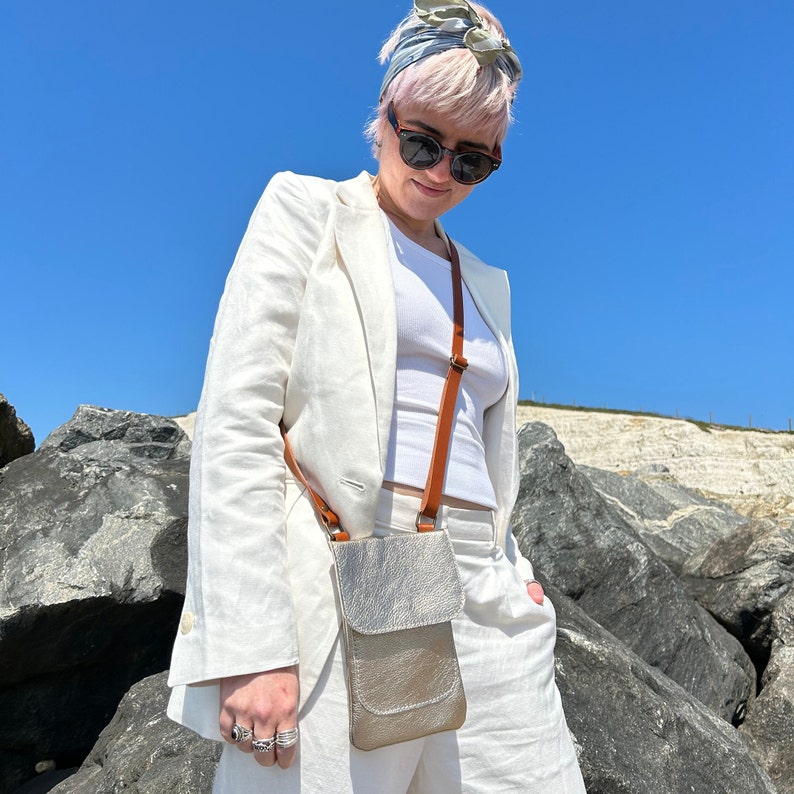 Girl with short blond hair wearing a white linen suit and a Champagne gold leather smartphone bag with a crossbody strap standing in front of a rocky landscape and a perfect blue sky