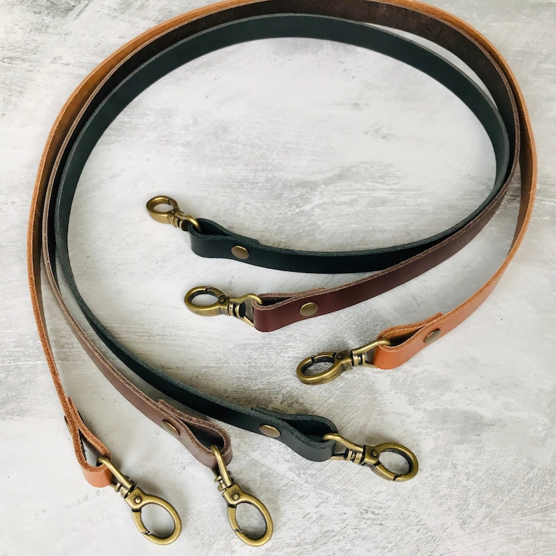 Three leather bag straps, black, brown and tan with antique brass lobster clasps and rivet detail sitting on a distressed grey background