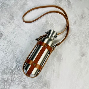 Tan cage style leather water bottle carrier with a crossbody strap and antique brass rivets holding a stainless steel water bottle