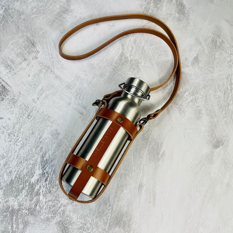 Tan cage style leather water bottle carrier with a crossbody strap and antique brass rivets holding a stainless steel water bottle