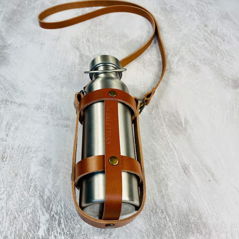 Tan cage style leather water bottle carrier with a crossbody strap and antique brass rivets holding a stainless steel water bottle