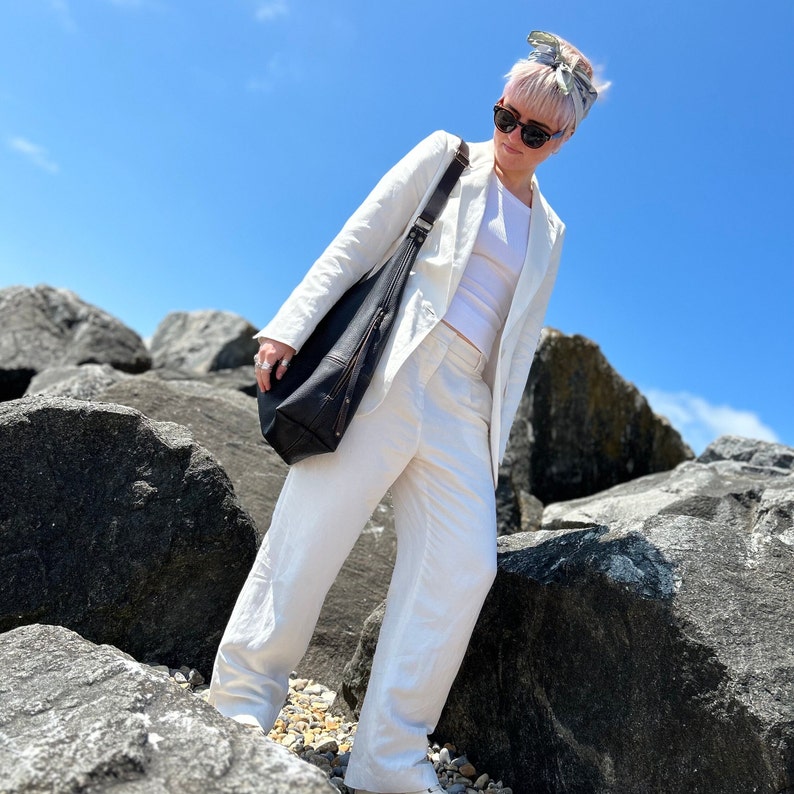 A girl with a bleach blonde pixie cut wearing a white linen suit, sunglasses and a large dark brown leather hobo bag with a cross body strap on a rocky beach with a perfect blue sky