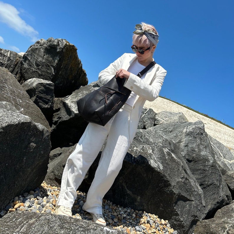 A girl with a bleach blonde pixie cut wearing a white linen suit, sunglasses and a large dark brown leather hobo bag with a cross body strap on a rocky beach with a perfect blue sky