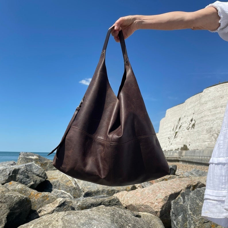 Girls arm holding up a large brown leather hobo bag on a rocky beach with a perfect blue sky