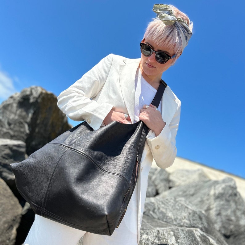 A girl with a bleach blonde pixie cut wearing a white linen suit, sunglasses and a large dark brown leather hobo bag with a cross body strap on a rocky beach with a perfect blue sky