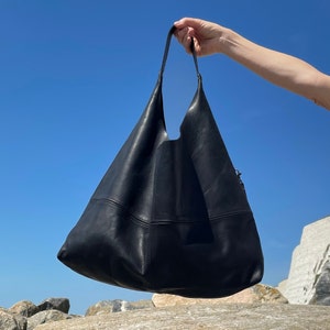 Girls arm holding up a large black leather hobo bag on a rocky beach with a perfect blue sky