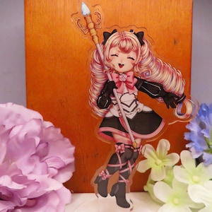 Puede incluir: Un personaje de estilo anime colorido con ropa rosa y negra, sosteniendo un bastón con una parte superior dorada y azul. El personaje tiene el pelo largo rubio con reflejos rosas y sonríe. El fondo es una superficie de madera.