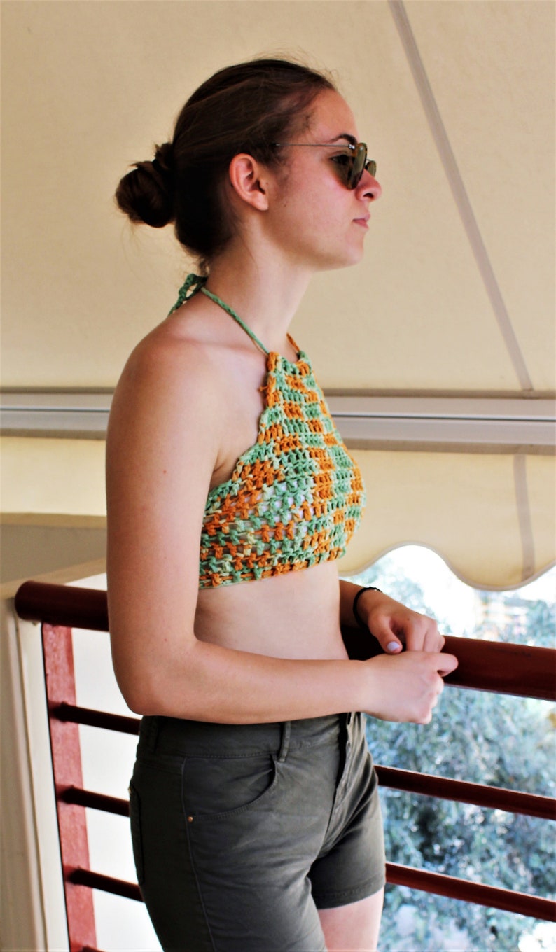 May include: A woman wearing a green and orange crochet halter top and dark green denim shorts. She is standing in front of a white awning.