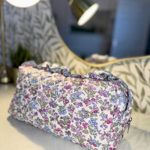 May include: A quilted cosmetic bag with a floral pattern in shades of pink, purple, and blue on a white background. The bag has a ruffled edge and a zipper closure. The bag is rectangular in shape.