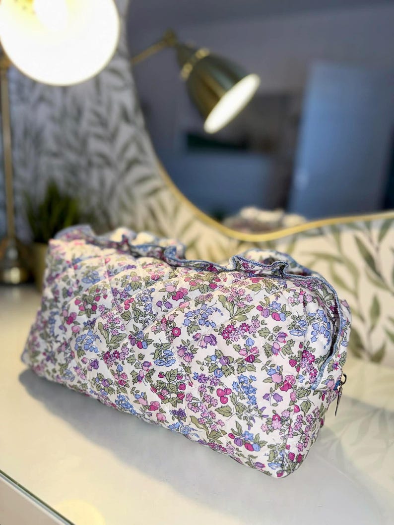 May include: A quilted cosmetic bag with a floral pattern in shades of pink, purple, and blue on a white background. The bag has a ruffled edge and a zipper closure. The bag is rectangular in shape.