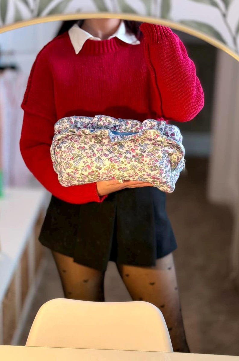 May include: A floral print clutch with a drawstring closure. The bag features a pattern of small pink, blue, and yellow flowers on a white background. The person is wearing a red sweater and black shorts.