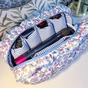 May include: Floral print cosmetic bag with a blue zipper. The bag contains a hair dryer, brushes, and other beauty tools. The bag is white with pink and purple flowers and a blue gingham interior.