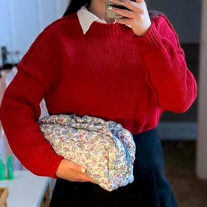 May include: A person wearing a red sweater over a white collared shirt and a black skirt. They are holding a floral print bag with a blue zipper. The outfit is completed with patterned tights.