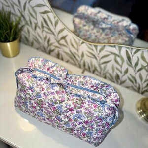 May include: A floral print cosmetic bag with a blue zipper and trim. The bag features a quilted design with a pattern of pink, blue, and green flowers on a white background. The bag is sitting on a white surface.