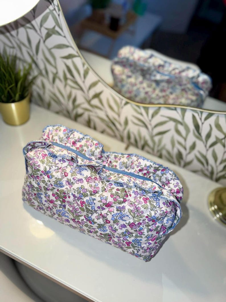 May include: A floral print cosmetic bag with a blue zipper and trim. The bag features a quilted design with a pattern of pink, blue, and green flowers on a white background. The bag is sitting on a white surface.