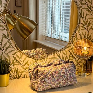 May include: A floral print quilted cosmetic bag with a ruffled edge sits on a vanity. The bag features a pattern of small flowers in pink, purple, and blue on a white background. A gold-toned lamp and mirror are in the background.