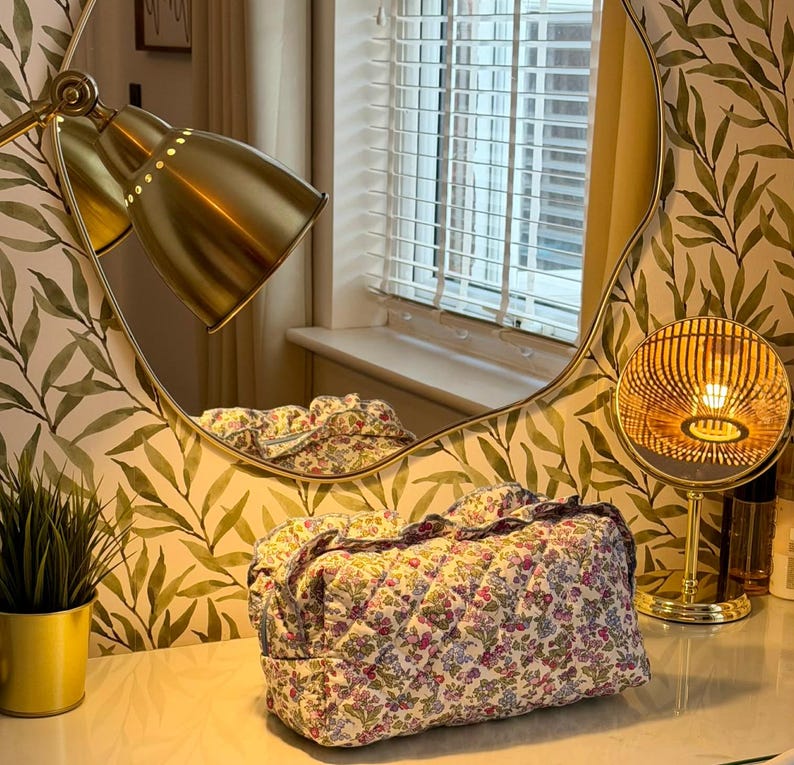 May include: A floral print quilted cosmetic bag with a ruffled edge sits on a vanity. The bag features a pattern of small flowers in pink, purple, and blue on a white background. A gold-toned lamp and mirror are in the background.