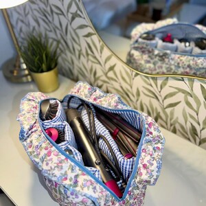 May include: A floral print cosmetic bag with a blue gingham interior and a blue zipper. The bag is open, revealing hair styling tools. The bag is sitting on a white surface, with a mirror in the background.