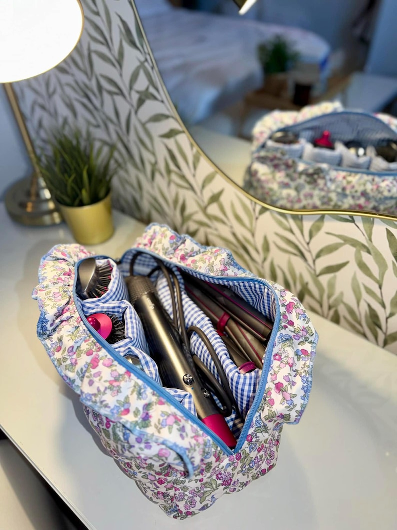 May include: A floral print cosmetic bag with a blue gingham interior and a blue zipper. The bag is open, revealing hair styling tools. The bag is sitting on a white surface, with a mirror in the background.