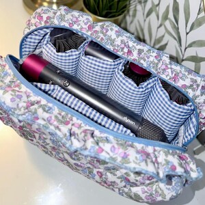 May include: A floral print travel case with a blue zipper and gingham interior. The case holds a Dyson hair dryer and several hair brushes. The case is white with a pattern of pink and purple flowers.