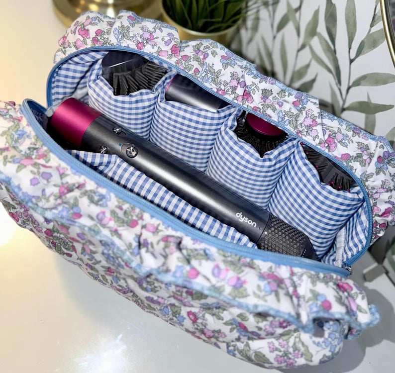 May include: A floral print travel case with a blue zipper and gingham interior. The case holds a Dyson hair dryer and several hair brushes. The case is white with a pattern of pink and purple flowers.