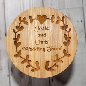 May include: A round wooden box with a floral design and the text "Jodie and Chris' Wedding Fund" engraved on the front.