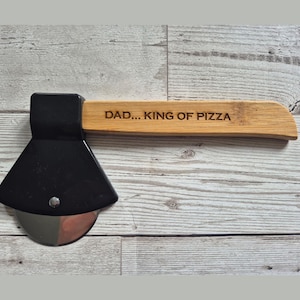 Può includere: Un taglia pizza in legno a forma di ascia con manico nero e lama argentata. Il manico è inciso con il testo "DAD...KING OF PIZZA".