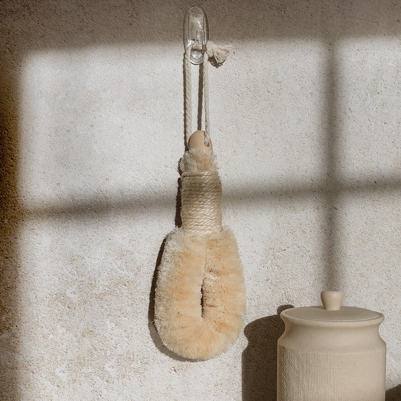 May include: A beige, oval-shaped body brush with stiff, natural-colored bristles and a wooden handle. The brush features a thick, twisted rope cord attached to the top for hanging, and the handle is wrapped in a matching rope texture.