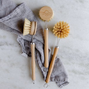 May include: Three wooden dish brushes with different bristle types. The brushes have wooden handles and metal wire loops. The top brush has a wooden head with a sun and waves design.