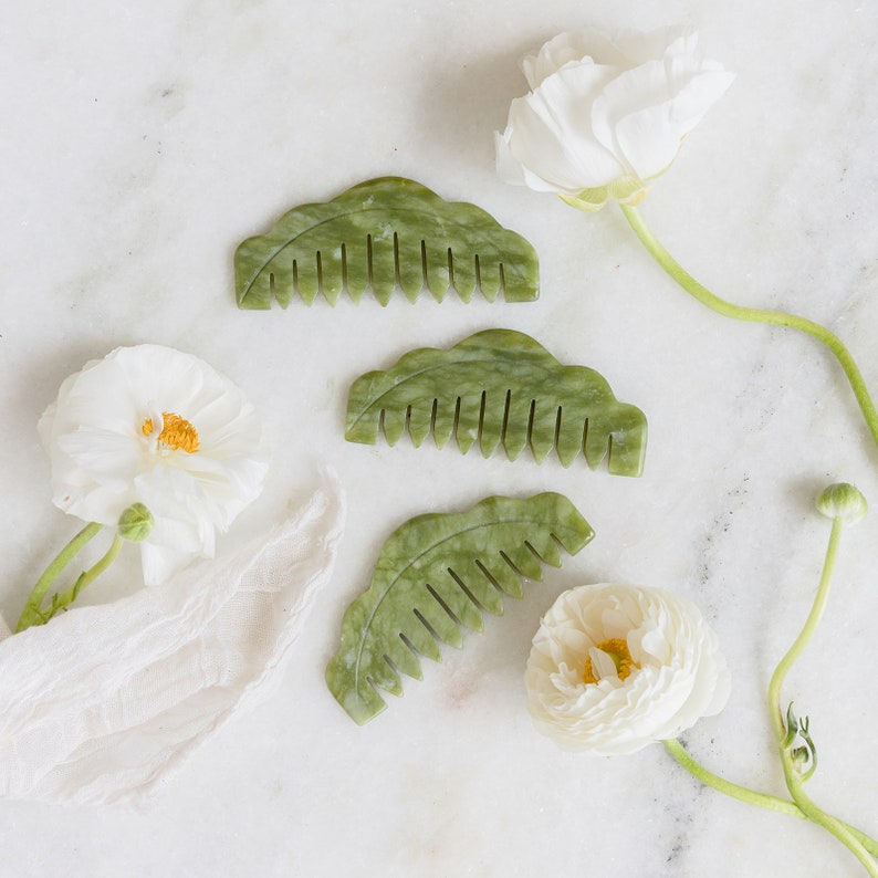 May include: Three green jade combs with a leaf-like design. The combs are on a white marble surface with white flowers.