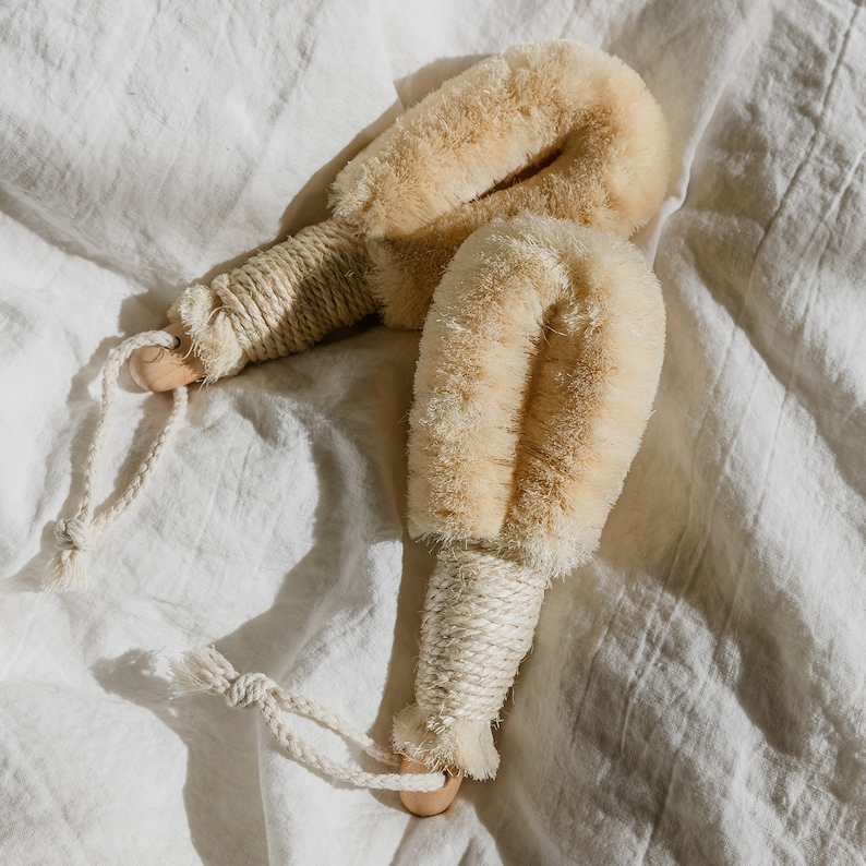 May include: Two bath brushes with light beige, stiff natural bristles arranged in a curved, elongated shape. Each brush features a wooden handle wrapped in thick, light-colored rope and includes a short, braided rope loop attached to the end.