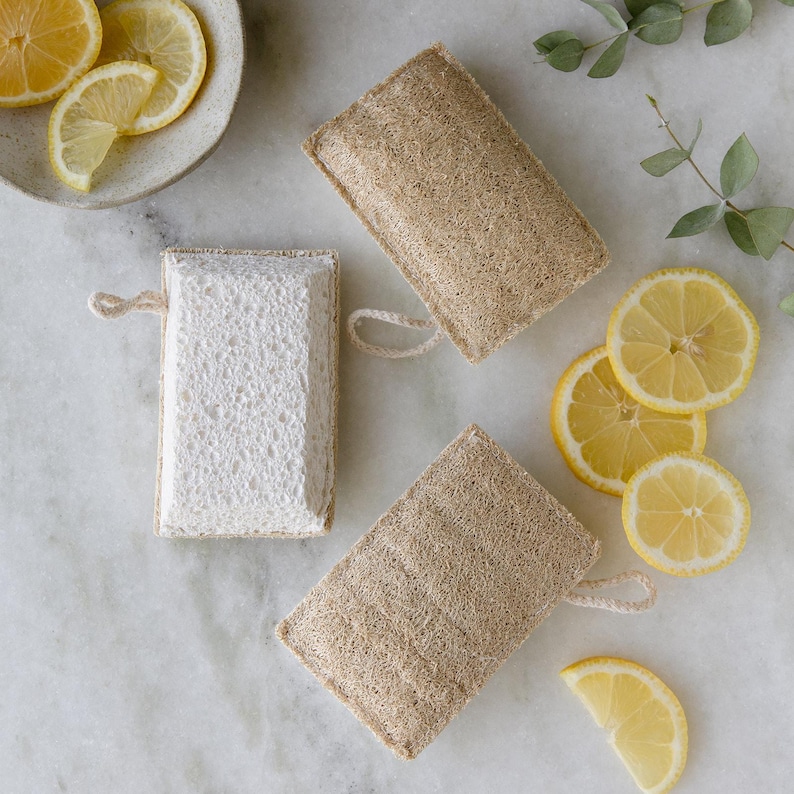 May include: Three natural loofah sponges, two are beige and one is white. The sponges are rectangular and have a string attached to one corner. There are lemon slices on a white surface.
