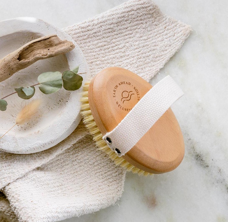 Puede incluir: Un cepillo corporal redondo de madera con cerdas naturales claras y una correa de mano de lona blanca. El mango de madera presenta un logotipo circular con el texto Earth Ahead Wellness y un icono de onda, asegurado con dos remaches de metal.