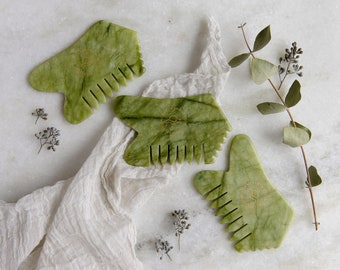 May include: Three green jade gua sha tools with gold accents and a comb-like design. The tools are on a white fabric background.