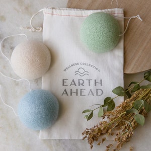May include: Three soft, textured cleansing sponges in a white drawstring bag with the text "Wellness Collection Earth Ahead". The sponges are beige, light blue, and light green.
