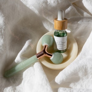 May include: A green jade facial roller with a rose gold handle sits in a white bowl with a bottle of green facial oil labeled "Blue Tansy Radiance".