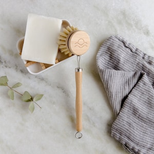 May include: A wooden dish brush with a natural bristle brush head and a wooden handle with a wave logo. The brush is next to a white bar of soap in a white dish.
