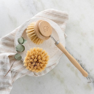 May include: A wooden dish brush with a long handle and a round brush head, alongside a smaller, round scrub brush. The brushes are made of light-colored wood and natural bristles. A white and beige striped cloth and a sprig of eucalyptus are also in the image.
