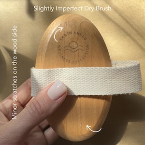Puede incluir: Un cepillo de madera ovalado para el cuerpo con una correa de tela tejida de color claro en el centro. La madera presenta el texto grabado Earth Ahead Wellness Collection con un icono de sol y olas. La superficie muestra peque&ntilde;os ara&ntilde;azos.