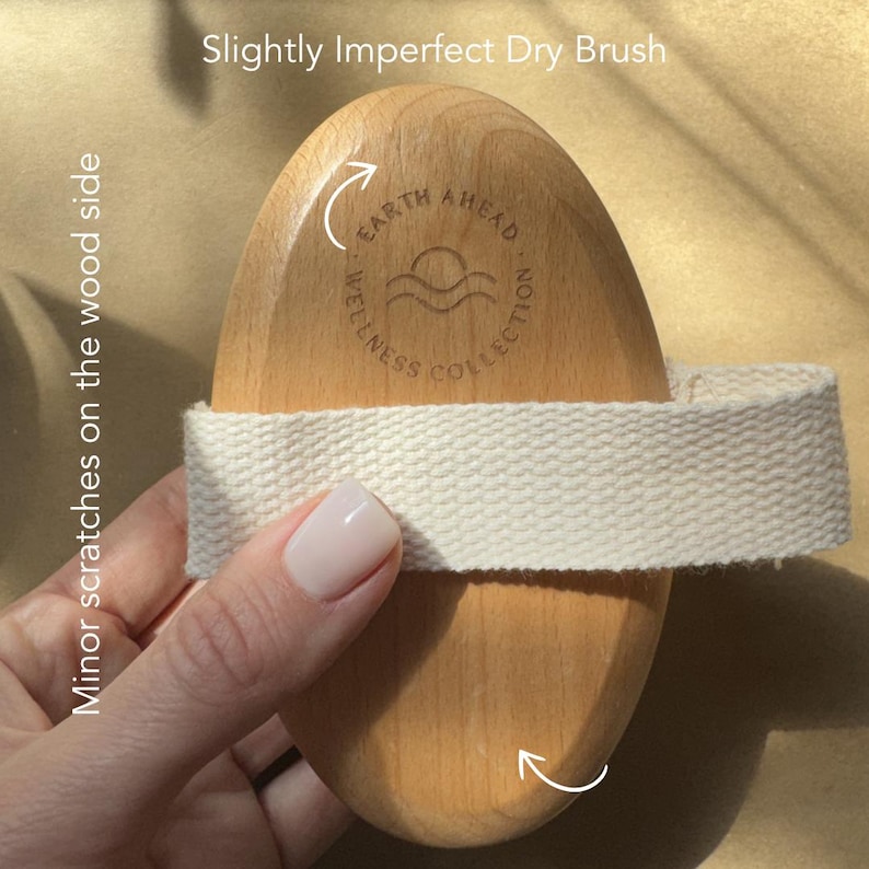 Puede incluir: Un cepillo de madera ovalado para el cuerpo con una correa de tela tejida de color claro en el centro. La madera presenta el texto grabado Earth Ahead Wellness Collection con un icono de sol y olas. La superficie muestra peque&ntilde;os ara&ntilde;azos.