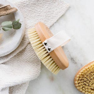 Puede incluir: Un cepillo corporal de forma ovalada con mango de madera clara y cerdas densas de color p&aacute;lido. Una correa ancha de lona color blanco roto est&aacute; fijada a la parte superior del mango de madera con dos remaches met&aacute;licos plateados para un agarre.
