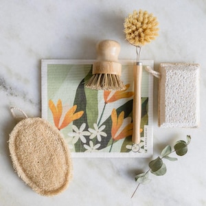 May include: A set of eco-friendly cleaning tools including a wooden handled brush with natural bristles, a loofah sponge, and a white sponge. The tools are arranged on a green and white patterned kitchen towel with the text "Earth Friendly" printed on it.