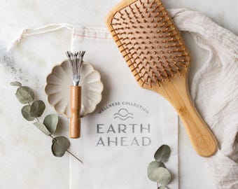 May include: A rectangular bamboo hairbrush with wooden bristles, a small metal hairbrush cleaner with a wooden handle, and a white cotton drawstring bag printed with the text WELLNESS COLLECTION EARTH AHEAD, accented by green eucalyptus leaves.