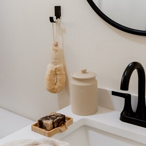 May include: A beige bath brush with a rope handle hangs from a black wall hook. Next to it sits a cylindrical beige canister with a lid and a rectangular bamboo soap dish holding a marbled brown and black bar of soap near a black faucet.