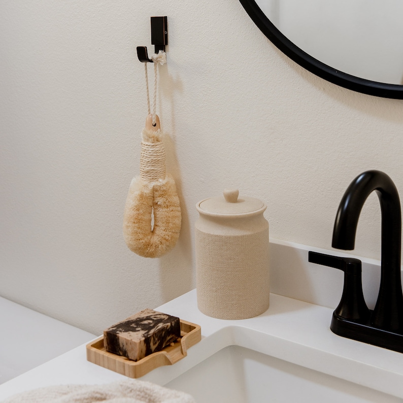 May include: A beige bath brush with a rope handle hangs from a black wall hook. Next to it sits a cylindrical beige canister with a lid and a rectangular bamboo soap dish holding a marbled brown and black bar of soap near a black faucet.