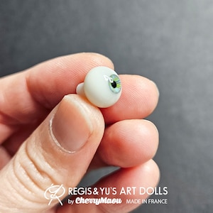 May include: Close-up of a single glass eye for doll making. The eye is white with a green iris and black pupil. The eye is being held by a hand.
